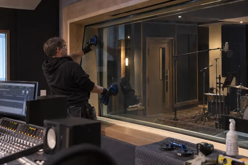 Recording studio interior from control desk: technician glazing a large angled acoustic observation screen within deep timber jambs, dark acoustic panels and microphone booth beyond.
