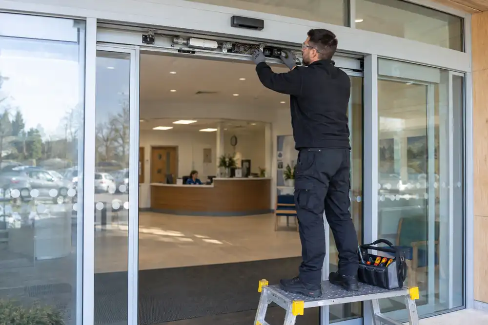 Modern clinic entrance: technician inspecting the overhead operator of automatic sliding glass doors from a secure low podium, doors parted centrally, clear visible aluminium frame and sensor.