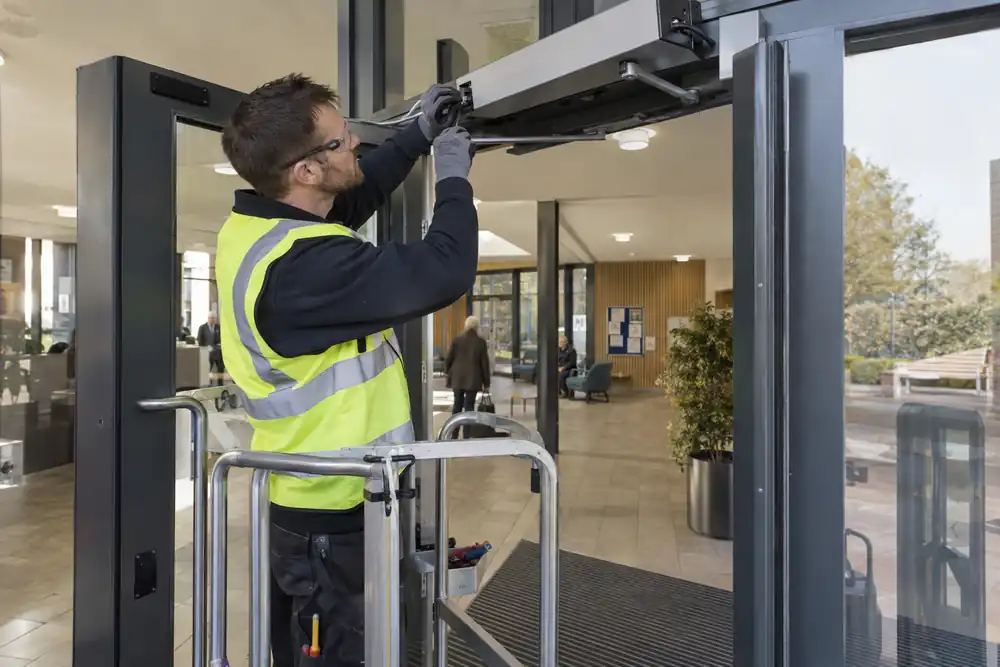 Community building lobby: technician adjusting the articulated overhead operator on an automatic swing glass door held partly open, safe podium with rail, oblique doorway view.
