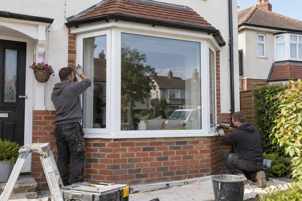 1930s semi-detached house front: installers finishing a newly formed ground-floor projecting bay window with three glazed facets, proper brick base and tiled canopy, wide daylight view.