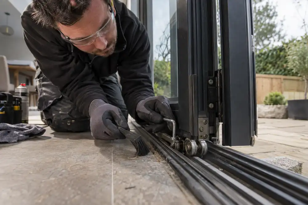 Macro low-angle trade photograph: kneeling technician servicing the bottom roller carriage and cleaning the track of partially folded patio bifold doors, small brush and hex key, glass visible above.