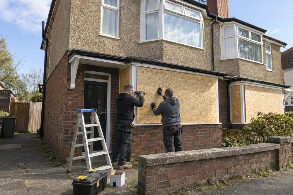 Wide view of a vacant two-storey British semi-detached house with ground-floor windows securely boarded; pair of tradespeople finishing one panel from pavement level, clear afternoon.