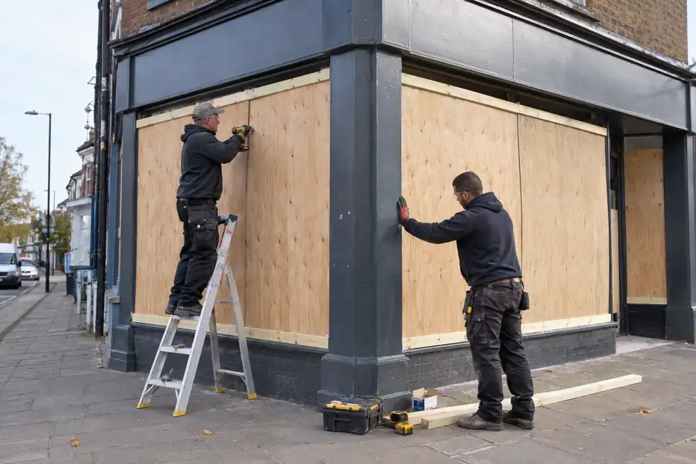 Corner high-street retail frontage with wide display window: two workers fixing large precut plywood sheets to continuous timber battens, daylight, bare blank fascia, glass debris already cleared.