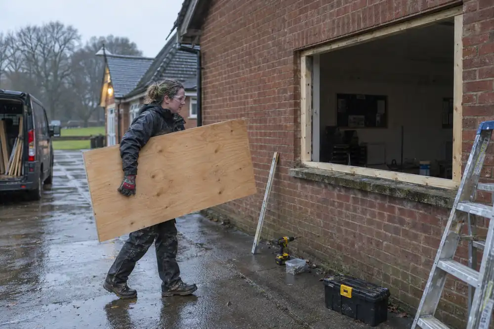 Rainy early morning at a small brick community hall: tradeswoman carrying a measured plywood board toward a fully cleared ground-floor window opening with timber battens prepared, wide side view.