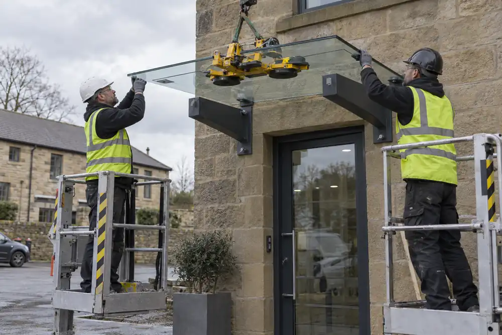 Exterior stone office doorway: two installers on guarded podium platforms positioning a small laminated glass canopy onto stout wall-mounted steel brackets using a portable glass lifter, ground visible, overcast daylight.