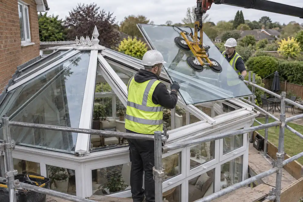 Garden conservatory refurbishment: team replacing roof glazing in white structural rafters using vacuum lifter, working from fully guarded access platforms alongside roof, no stepping onto glass, exterior elevated view.