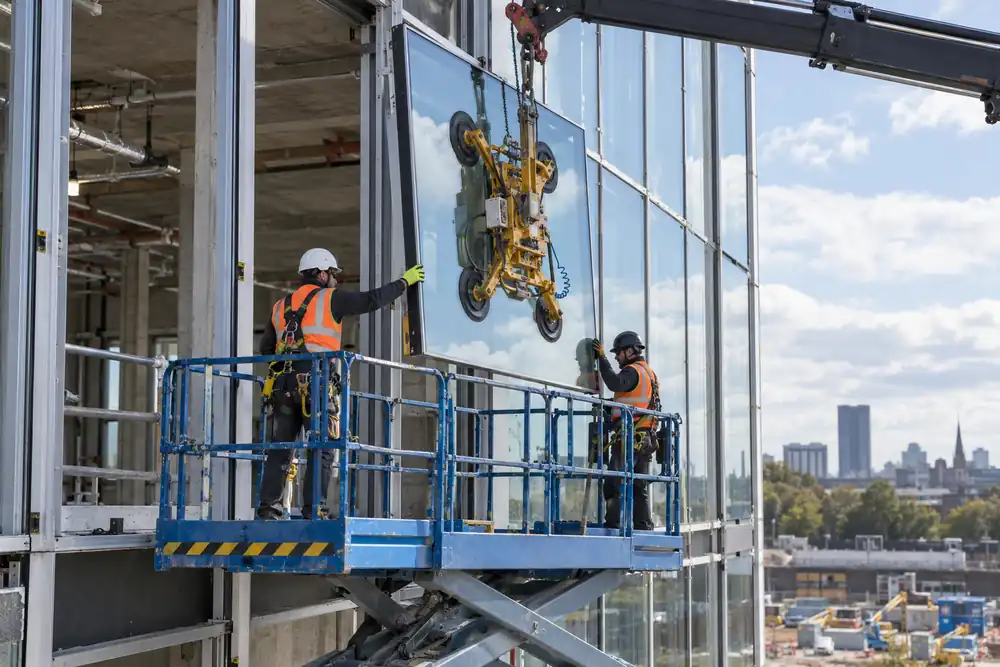 Office building facade under construction: installers on fully guarded scissor lift guide a rectangular glazed curtain-wall panel into aluminium mullions with crane-mounted vacuum lifter, wide exterior daylight view.