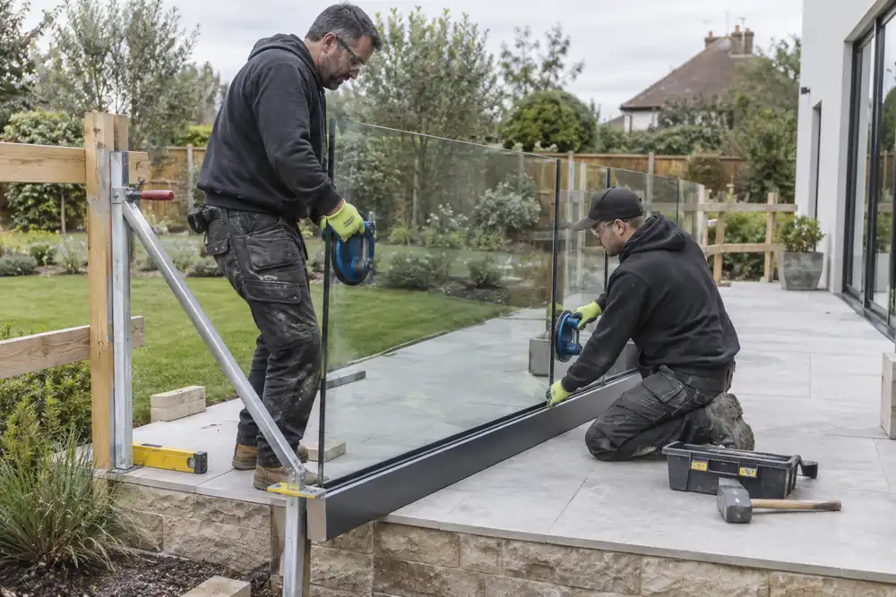 Ground-floor raised terrace: glaziers seat a clear laminated glass balustrade panel into a continuous metal base channel, suction lifters and sturdy supports, temporary edge barrier, landscaped garden backdrop.