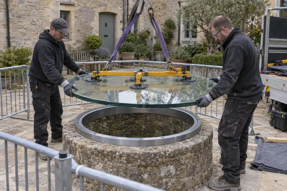 Stone courtyard with an old circular well: two installers lower a thick circular laminated glass cover onto a continuous engineered steel support ring using a lifting rig, perimeter barrier, nobody standing on cover.